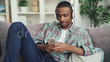 漂亮的非裔美国学生正在使用智能手机触摸屏幕，在沙发上用耳机听音乐
